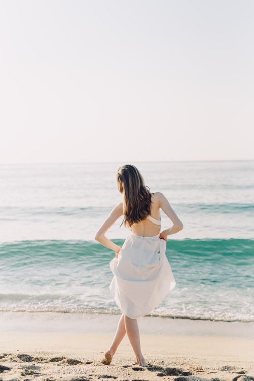 Woman in a flowing white linen dress walking barefoot on the beach, coastal chic festival style